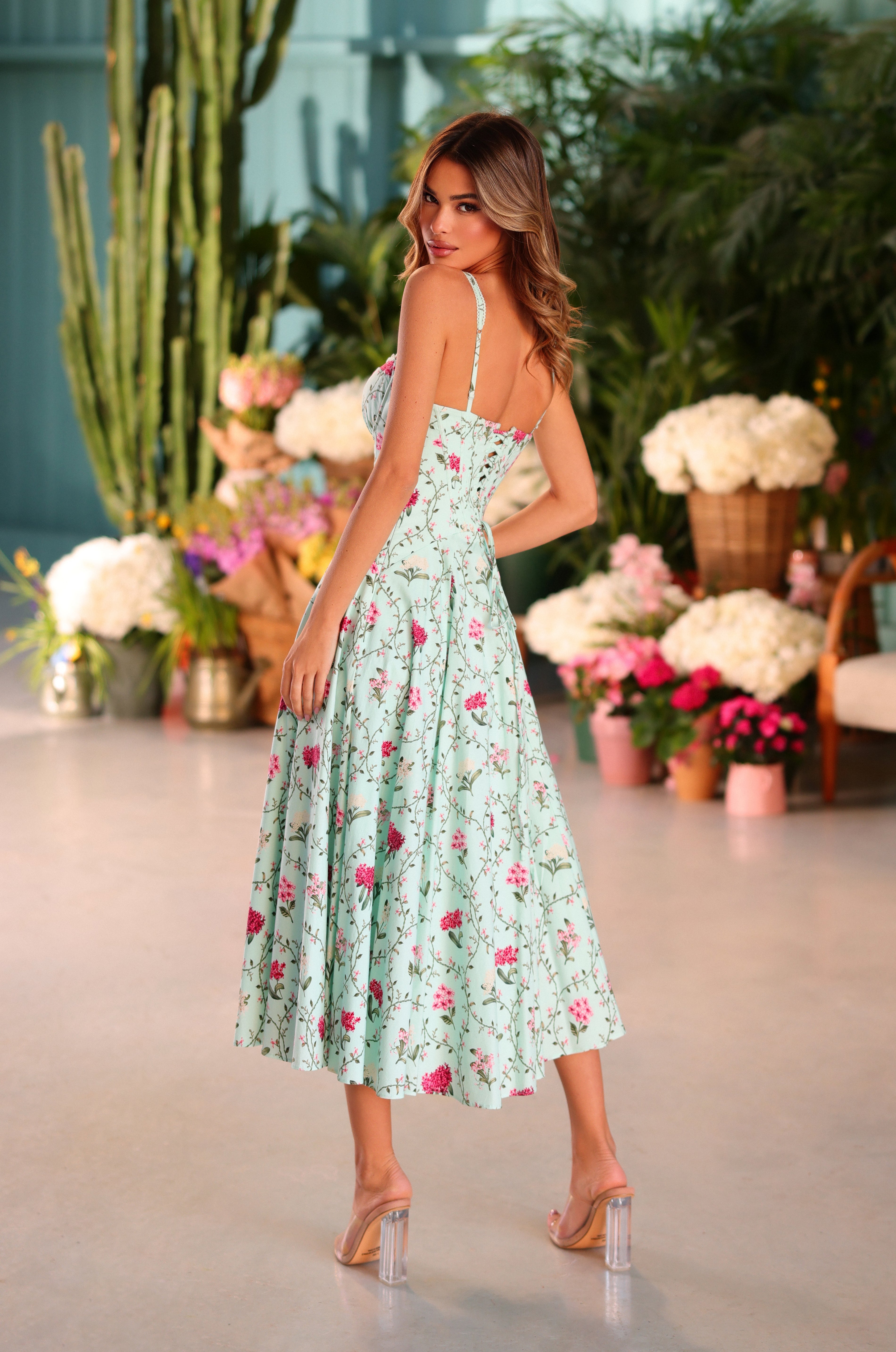 A woman in the Rosella Mint Green Floral Casual Midi Dress with a corset bodice and clear heels stands in a bright room filled with potted plants and flowers.